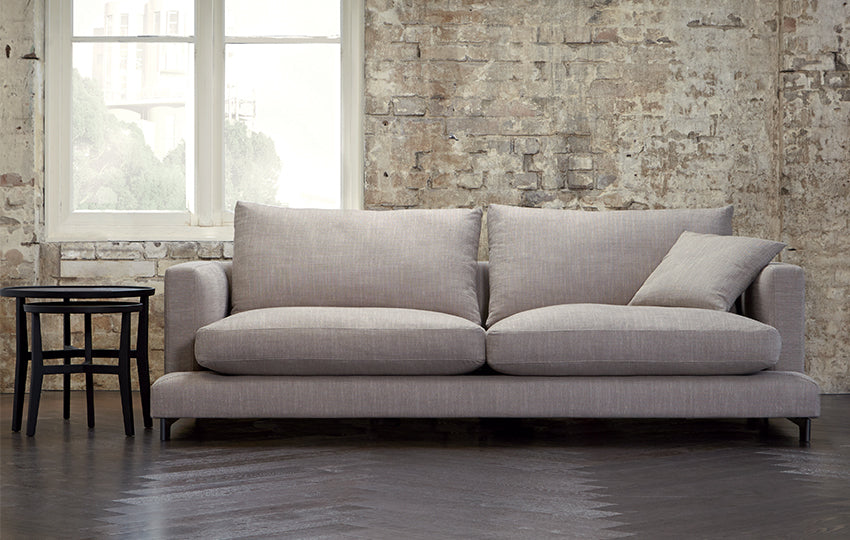 The Lazy Sofa in a room with exposed brick a Window showing to the outside and a nest of tables. 